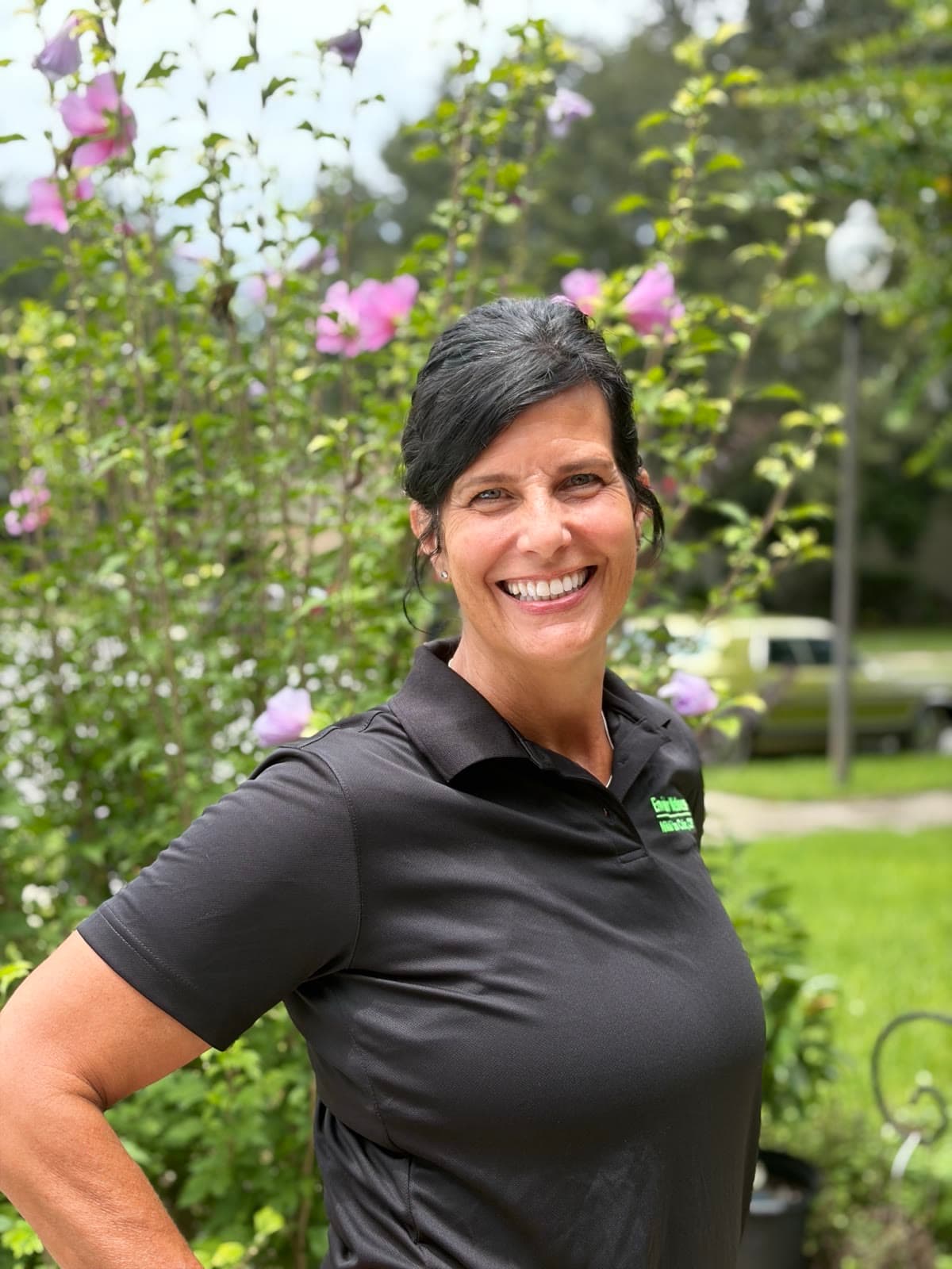 Nikki Van Culin standing outdoors in warm light, hand on hip, smiling, flowering shrubs behind her